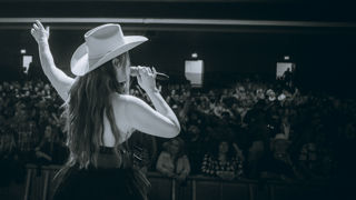 A female member of the country music band One Night in Nashville stands with her back to the camera, facing a packed crowd. She has one hand lifted in the air while the other holds a microphone which she is singing into, wearing a cowboy hat.