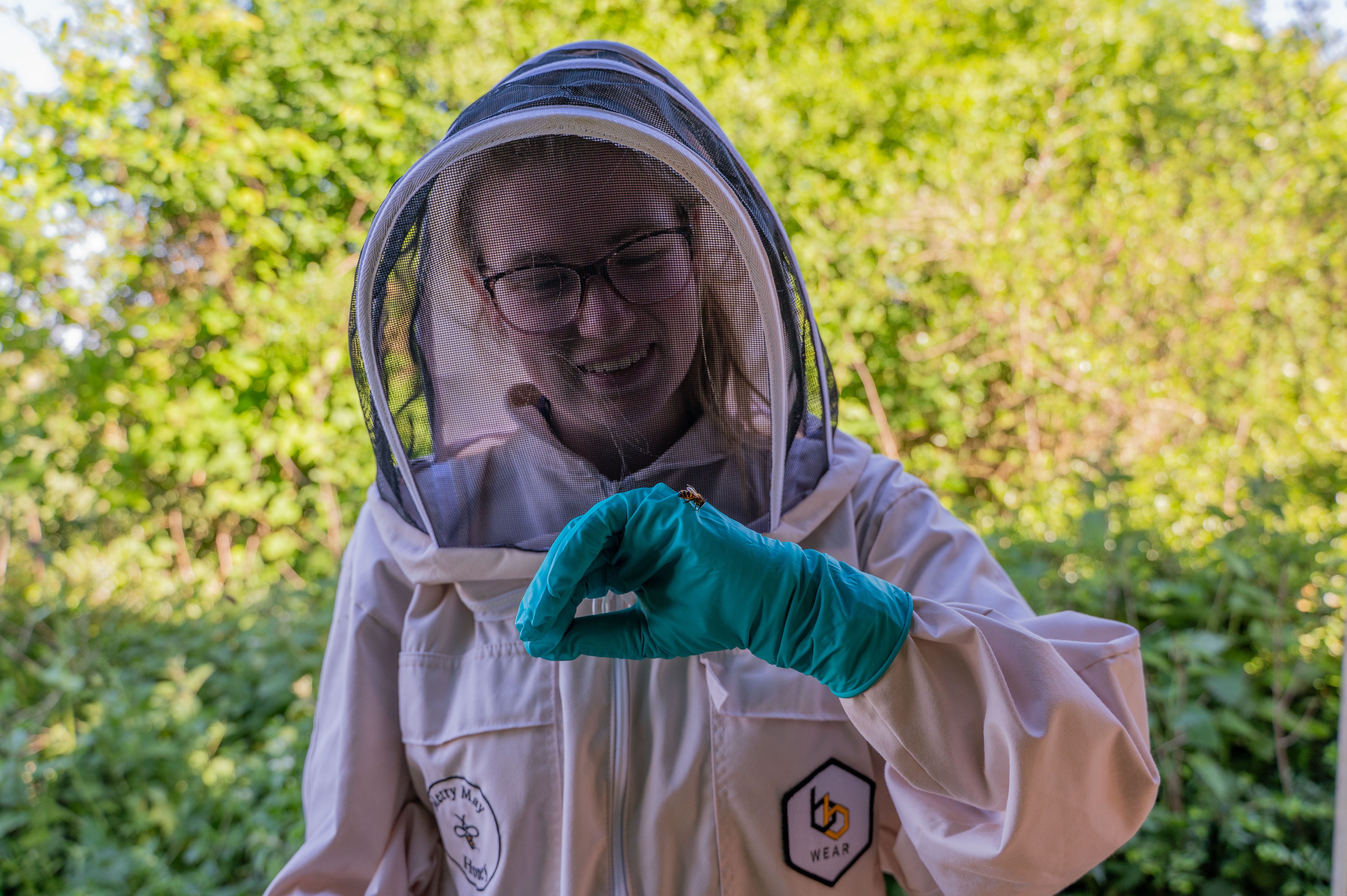 Jade from Cotswold Farm Park team wears a big white bee suit and helmet, holding up one hand with a bee resting on it. 
