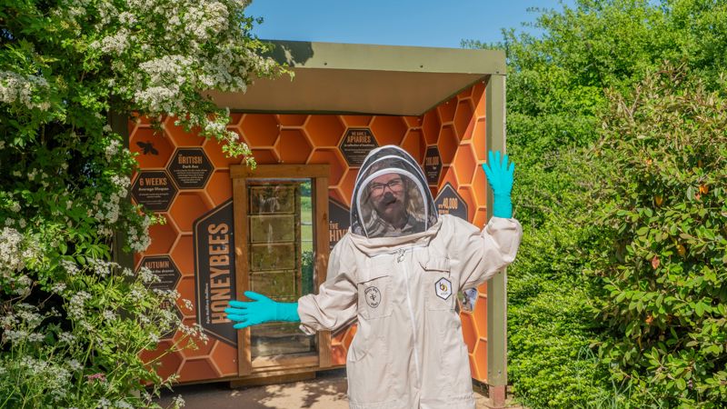 Jade, the Cotswold Farm PArk trainee beekeeper stands in front of a bee hive display surrounded by tall bushes in the sunshine. She is wearing a white bee suit and helmet and is smiling and waving her hands at the camera.