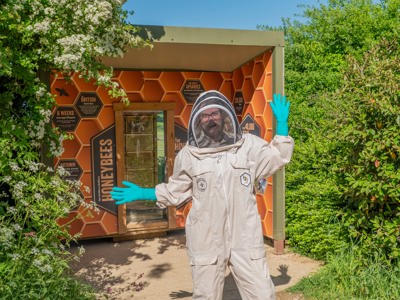 Jade, the Cotswold Farm PArk trainee beekeeper stands in front of a bee hive display surrounded by tall bushes in the sunshine. She is wearing a white bee suit and helmet and is smiling and waving her hands at the camera. 