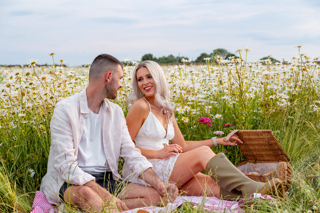 A man and woman wearing summer clothing sit in a field of beautiful flowers on a pink gingham picnic blanket having a romantic picnic for two in the Cotswold Farm Park Flower Fields. A brown wicker basket is open next to them, with tasty goodies inside.