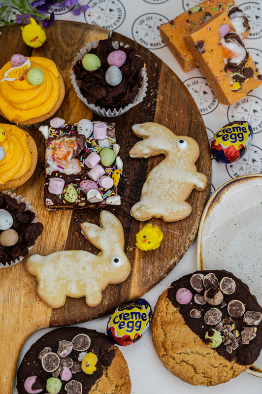 Easter baking laid out on a wooden platter. Includes creme eggs, rabbit shaped shortbread, cookies half dipped in chocolate with mini eggs and chocolate cornflake nests.