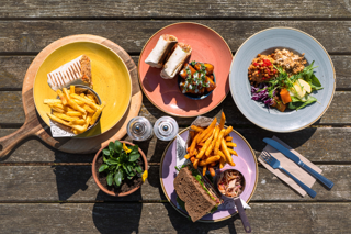 A picnic bench at the Cotswold Farm Park Ox Shed Restaurant from above with a spread of plates. Plates are yellow, red, blue and purple and show a range of items including hot wraps, chips, salads and a premium salt beef sandwich.