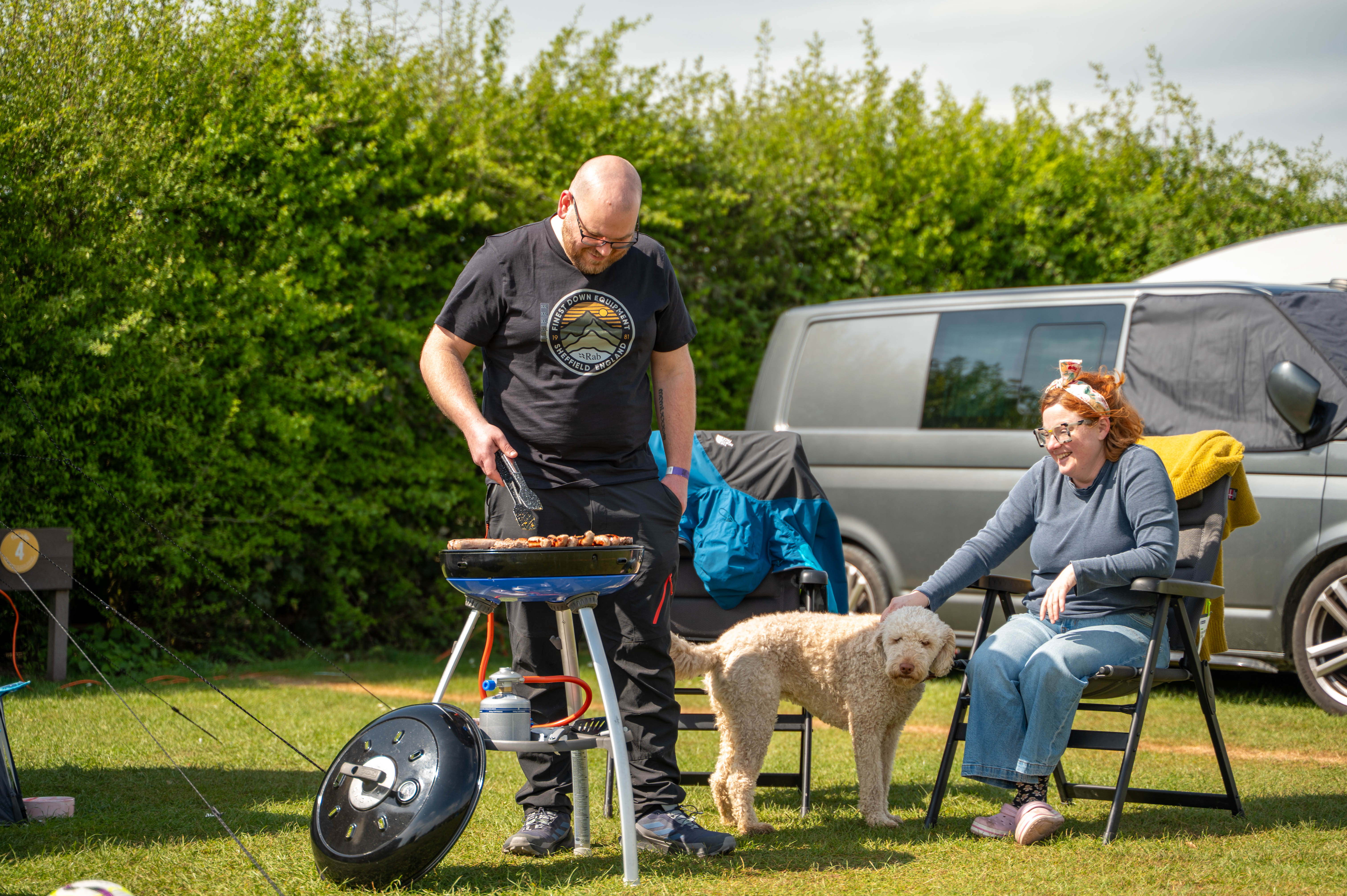 A man stands over a barbeque cooking while his wife watches on from a camping chair with their labradoodle in front of their converted van camper.