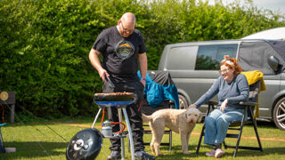 A man stands over a barbeque cooking while his wife watches on from a camping chair with their labradoodle in front of their converted van camper.
