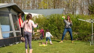 A lady plays a ball game on grass with her two young girls at Cotswold Farm Park Holidays on a sunny Easter weekend, while their grandparent watches on from inside a tent.
