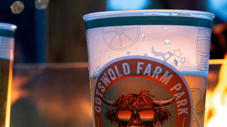 A pint of golden beer in a Cotswold Farm Park reusable cup in front of a barbeque