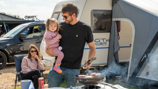 A dad stands cooking a barbeque outside his caravan at Cotswold Farm Park Holidays with his little girl on his hip with mum watching on.